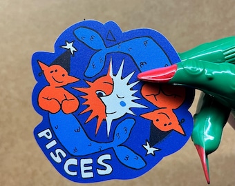May include: A die-cut sticker featuring a blue background with two orange fish figures, a central sun and moon graphic, and the word PISCES printed in white capital letters. The design includes small black star accents and black outlines.