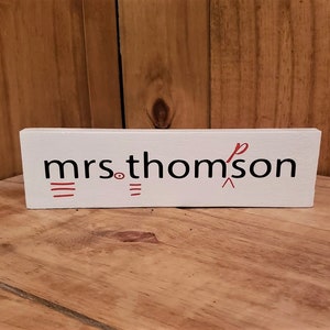 May include: A white wooden block with the name "mrs. thomson" printed in black and red. The block is rectangular and sits on a wooden surface. The text is stylized with red lines and a red symbol.