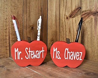 May include: Two red apple-shaped pen holders with white text that reads "Mr. Stewart" and "Ms. Chavez." Each holder has a brown stem and holds a pen. The background is a wooden surface.