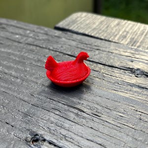 May include: A small, bright red figurine of a hen sitting in a nest. The object features a textured surface with visible horizontal lines, a rounded base, and a detailed shape representing a bird with a comb and tail feathers in a seated position.