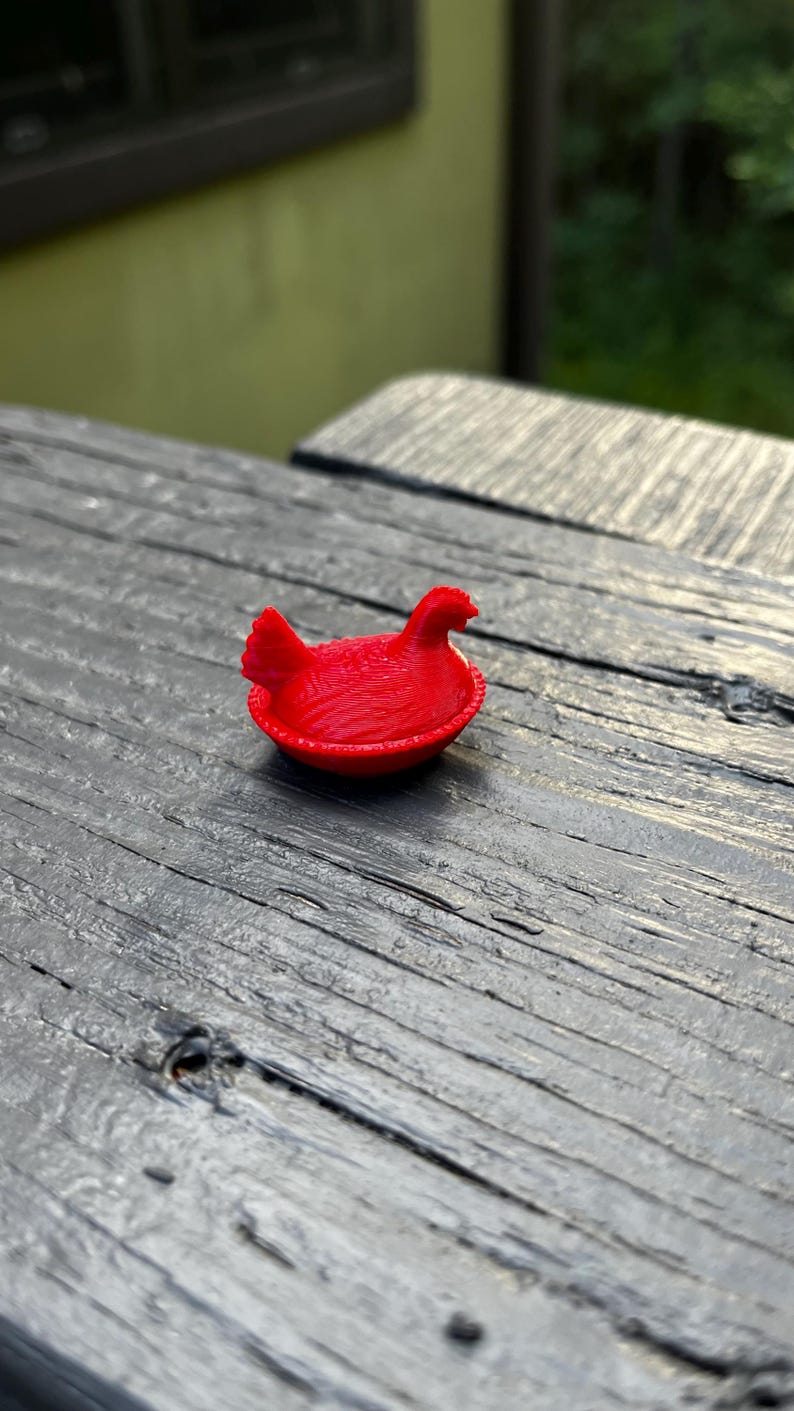 May include: A small, bright red figurine of a hen sitting in a nest. The object features a textured surface with visible horizontal lines, a rounded base, and a detailed shape representing a bird with a comb and tail feathers in a seated position.