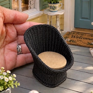 May include: A miniature black woven wicker chair featuring a rounded backrest and a circular tan seat cushion. The chair is small enough to be held in the palm of a hand, showcasing a detailed texture and a curved, open-front design for dollhouse use.