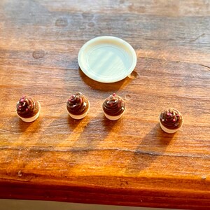 May include: A collection of miniature chocolate cupcakes with white frosting and pink sprinkles, alongside a small, round, white plate, on a wooden surface. The cupcakes have a glossy, dark brown appearance.