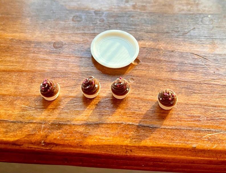 May include: A collection of miniature chocolate cupcakes with white frosting and pink sprinkles, alongside a small, round, white plate, on a wooden surface. The cupcakes have a glossy, dark brown appearance.