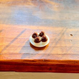 May include: A small white plate with four miniature cupcakes. Each cupcake has a white base and a dark chocolate frosting swirl, topped with a small decorative sprinkle. The plate is on a wooden surface.