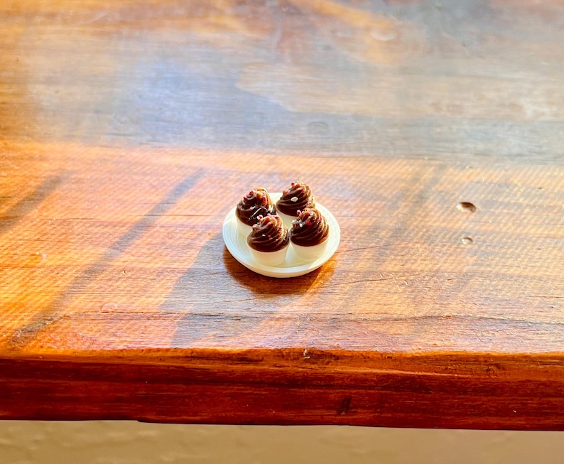 May include: A small white plate with four miniature cupcakes. Each cupcake has a white base and a dark chocolate frosting swirl, topped with a small decorative sprinkle. The plate is on a wooden surface.