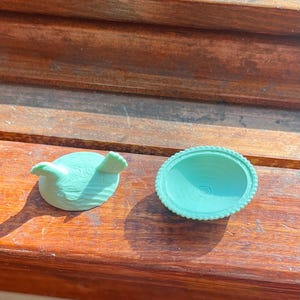 May include: A light mint green bird-shaped lid and a matching circular bowl with a beaded rim. Both items feature a textured surface and are rendered in a solid, matte mint green color, displaying a distinct layered finish from the manufacturing process.