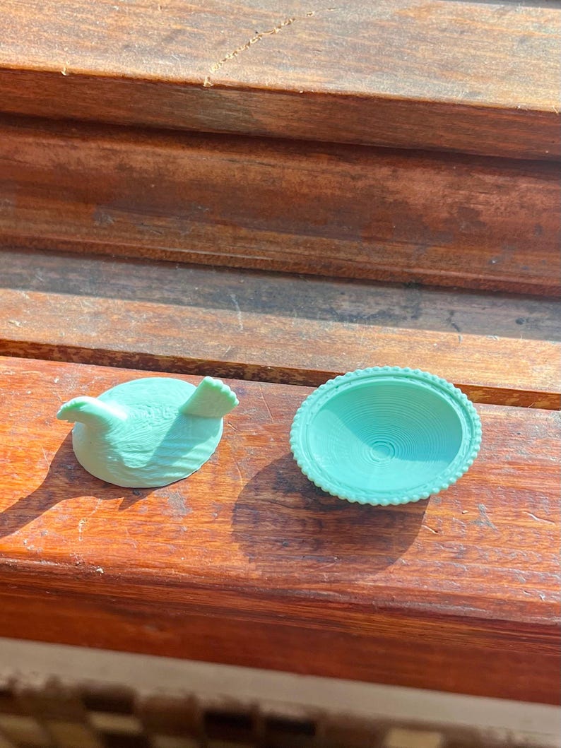 May include: A light mint green bird-shaped lid and a matching circular bowl with a beaded rim. Both items feature a textured surface and are rendered in a solid, matte mint green color, displaying a distinct layered finish from the manufacturing process.