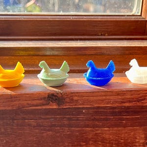 May include: Four small chicken figurines, each sitting in a nest-shaped base. The figurines are colored yellow, light green, blue, and translucent white. Each chicken has a textured surface representing feathers and is positioned in a seated posture.