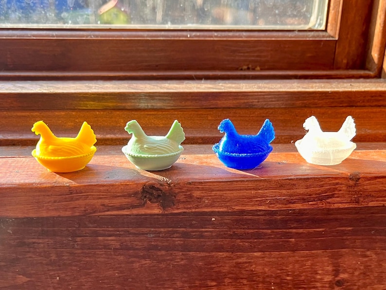 May include: Four small chicken figurines, each sitting in a nest-shaped base. The figurines are colored yellow, light green, blue, and translucent white. Each chicken has a textured surface representing feathers and is positioned in a seated posture.