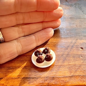 May include: A small white plate with four miniature chocolate desserts. Each dessert features a white base, topped with a swirl of dark chocolate frosting and white sprinkles. The plate rests on a wooden surface.