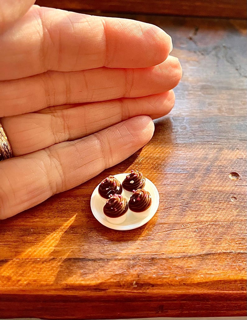 May include: A small white plate with four miniature chocolate desserts. Each dessert features a white base, topped with a swirl of dark chocolate frosting and white sprinkles. The plate rests on a wooden surface.