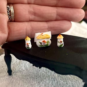 May include: Miniature ceramic mushroom-themed decor set. The set includes two small mushroom-shaped containers and a larger container with a lid, all painted with colorful details. The items are arranged on a small black table.