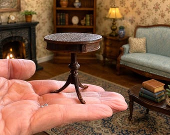 Może przedstawiać: Miniaturowy stolik na jednej nodze z ciemnobrązowego drewna z okrągłym blatem i trójnożną podstawą. Stolik posiada centralną kolumnę z ozdobnymi rzeźbieniami oraz trzy zakrzywione nogi, zaprojektowany do domku dla lalek, trzymany w dłoni.