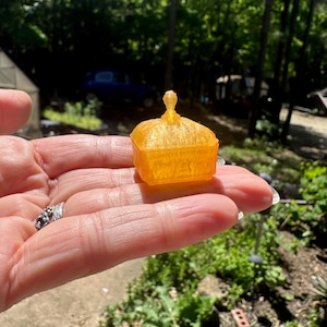 May include: A small, translucent orange-colored box with a lid, held in a hand. The box has a square shape with a decorative finial on top. The background shows a blurred outdoor scene with trees and greenery.
