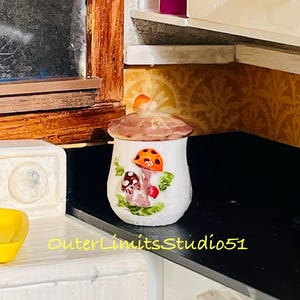 May include: A small white ceramic jar with a lid, featuring a painted design of orange and brown mushrooms with green leaves. The jar has a rounded shape and a small mushroom-shaped handle on the lid. The text OuterLimitsStudio51 is visible.