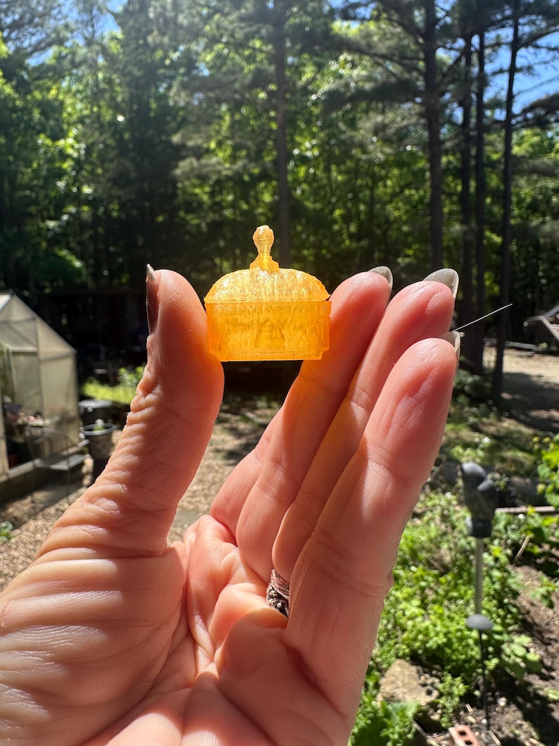 K&ouml;nnte beinhalten: Eine orangefarbene, durchscheinende Miniatur-Schatulle mit gew&ouml;lbtem Deckel und einem kleinen, zentralen knauff&ouml;rmigen Griff. Das Objekt hat eine texturierte Oberfl&auml;che und einen rechteckigen Boden, &auml;hnlich einem kleinen Dekogef&auml;&szlig; oder Spielzubeh&ouml;r.
