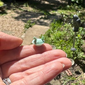 May include: A miniature light green figurine of a bird sitting inside a small white bowl. The bird has textured wings and a small beak, while the bowl has a smooth, rounded base. The entire object is small enough to rest on a fingertip.