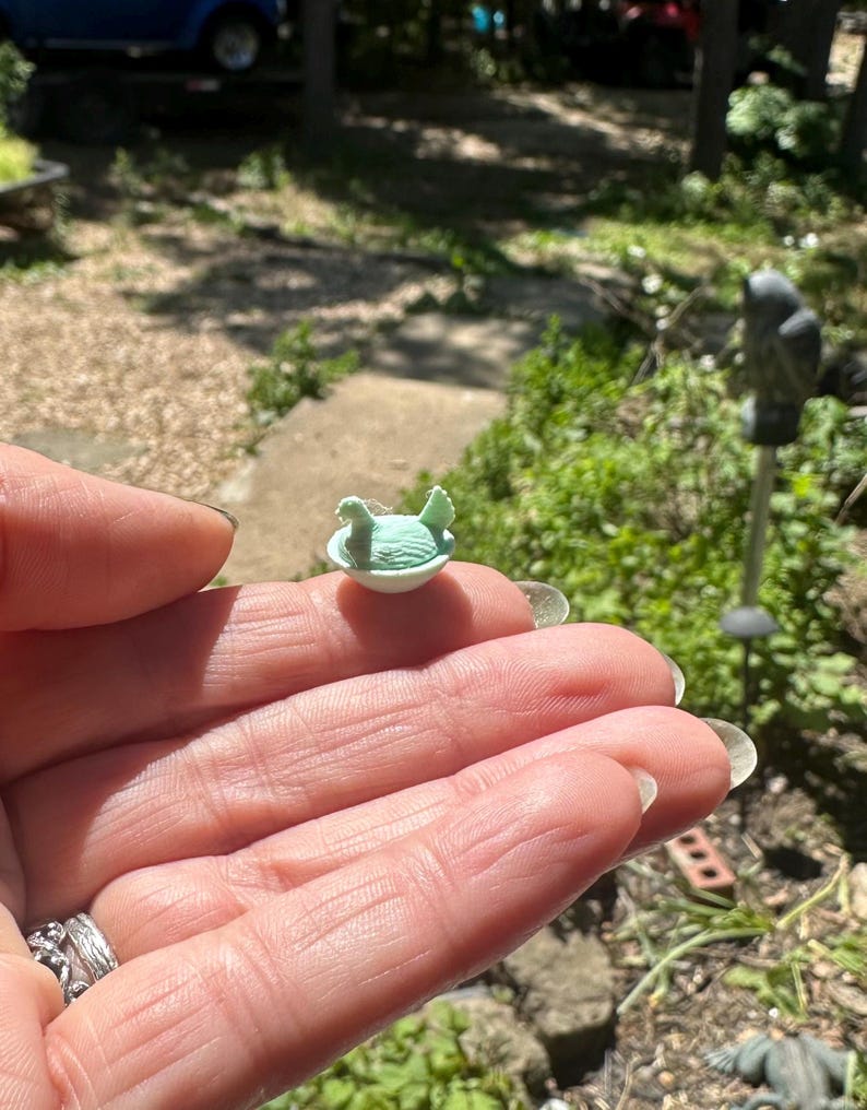 May include: A miniature light green figurine of a bird sitting inside a small white bowl. The bird has textured wings and a small beak, while the bowl has a smooth, rounded base. The entire object is small enough to rest on a fingertip.