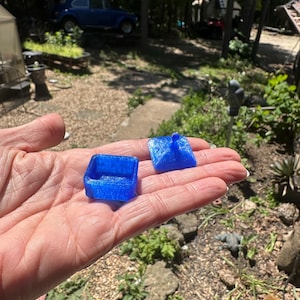 K&ouml;nnte beinhalten: Ein kleiner, quadratischer, durchscheinender blauer Beh&auml;lter mit passendem Deckel, der einen zentralen Knopf aufweist. Das Objekt hat eine texturierte, leicht glitzernde Oberfl&auml;che und ist in zwei Teilen dargestellt, bestehend aus dem offenen Boden und dem abnehmbaren Deckel.