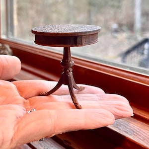 May include: A miniature brown table with a round top and three legs. The table has a detailed design and is held in a person's hand. The table is a decorative item.