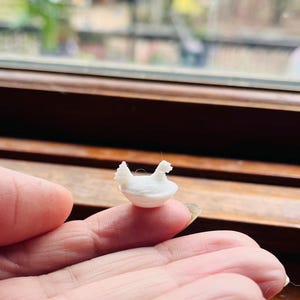 May include: A small, white, 3D-printed chicken figurine. The miniature sculpture features a detailed design with a bowl-like base and two chicken heads. The figurine is held between fingers, with a blurred window in the background.