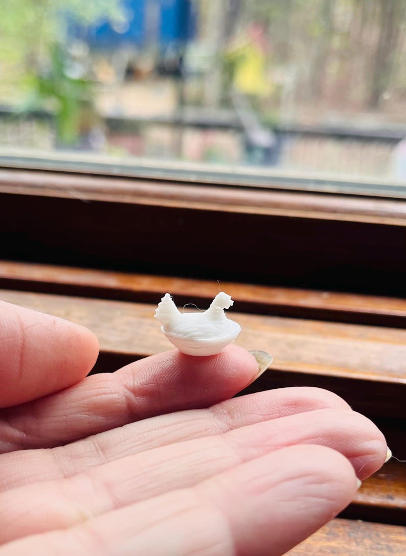 May include: A miniature white 3D printed figurine of a chicken sitting in a small bowl. The tiny object is rendered in a solid white material with visible horizontal layer lines from the printing process, showing a detailed bird shape.