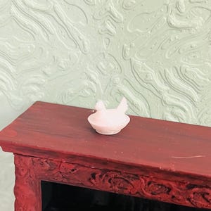 May include: A small, pale pink miniature figurine of a chicken sitting in a nest. The figurine is positioned on the flat, dark reddish-brown top surface of a piece of furniture with ornate, carved detailing along its front edge.
