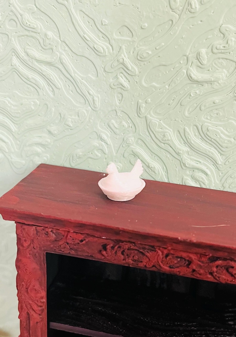 May include: A small, pale pink miniature figurine of a chicken sitting in a nest. The figurine is positioned on the flat, dark reddish-brown top surface of a piece of furniture with ornate, carved detailing along its front edge.