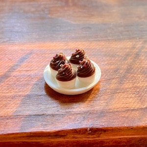 May include: Four miniature cupcakes with chocolate frosting and sprinkles on a small white plate. The cupcakes have white bases and are arranged on a white, oval-shaped plate. The image is taken on a wooden surface.