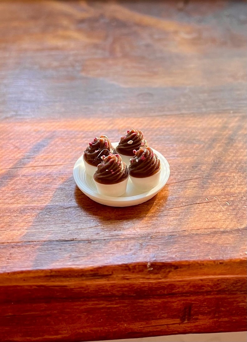 May include: Four miniature cupcakes with chocolate frosting and sprinkles on a small white plate. The cupcakes have white bases and are arranged on a white, oval-shaped plate. The image is taken on a wooden surface.
