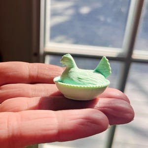May include: A miniature light green chicken figurine resting inside a small, white, round bowl with a textured, beaded rim. The chicken is depicted in a seated position with its head turned to the side and its tail feathers pointing upward.