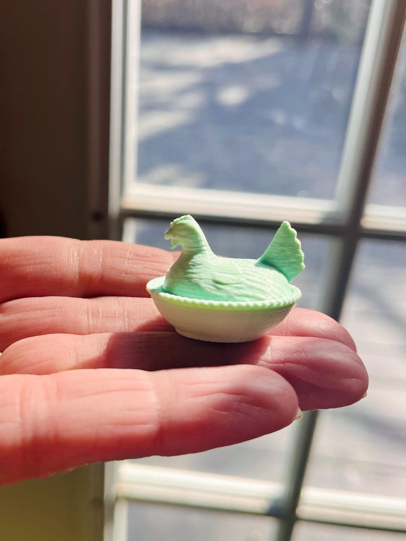 May include: A miniature light green chicken figurine resting inside a small, white, round bowl with a textured, beaded rim. The chicken is depicted in a seated position with its head turned to the side and its tail feathers pointing upward.