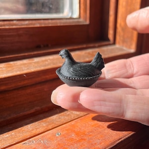 May include: A small, black, three-dimensional figurine of a hen sitting in a nest. The hen has a textured surface resembling feathers, and the nest features a beaded rim detail. The object is compact and shaped like a miniature lidded container.