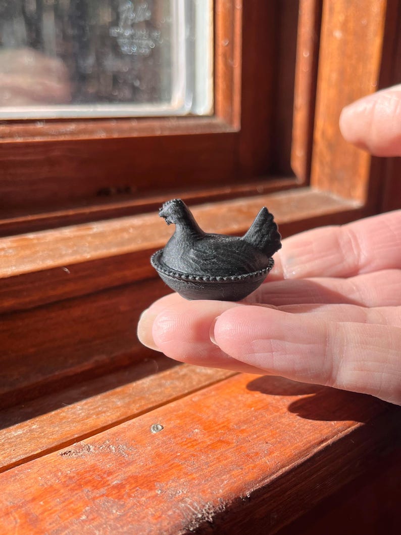 May include: A small, black, three-dimensional figurine of a hen sitting in a nest. The hen has a textured surface resembling feathers, and the nest features a beaded rim detail. The object is compact and shaped like a miniature lidded container.