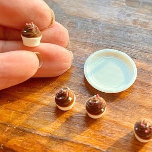 May include: Miniature chocolate cupcakes with white frosting and sprinkles. A small white plate is next to the cupcakes. The cupcakes are on a wooden surface.