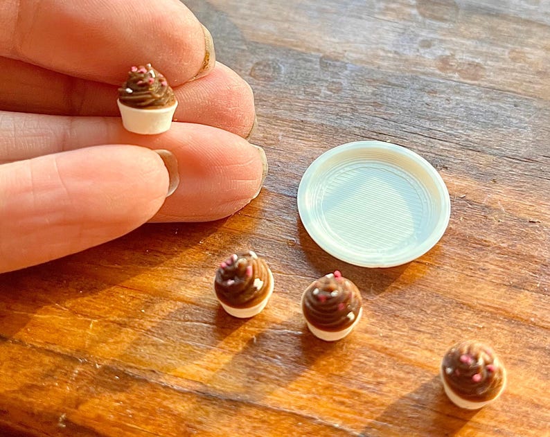 May include: Miniature chocolate cupcakes with white frosting and sprinkles. A small white plate is next to the cupcakes. The cupcakes are on a wooden surface.