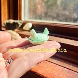 May include: A miniature light green glass figurine of a hen sitting in a nest, featuring a textured surface with detailed feathers and a basket-weave pattern on the base. The text OuterLimitsStudio51 is superimposed in yellow across the image.