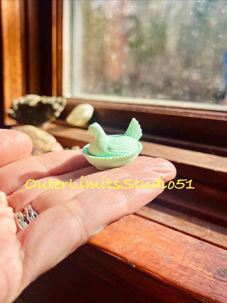 May include: A miniature light green glass figurine of a hen sitting in a nest, featuring a textured surface with detailed feathers and a basket-weave pattern on the base. The text OuterLimitsStudio51 is superimposed in yellow across the image.