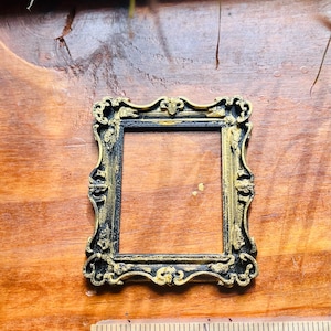 May include: Ornate, rectangular picture frame with a black and gold finish. The frame has a detailed, vintage design with decorative flourishes. The frame is approximately 2 inches wide and sits on a wooden surface.