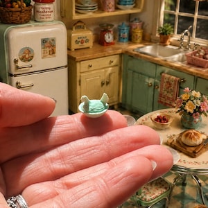 May include: A miniature light blue ceramic hen sitting in a small white bowl. The figurine features a textured surface representing feathers and a distinct head and tail shape. The item is held in a palm against a background of a dollhouse kitchen.