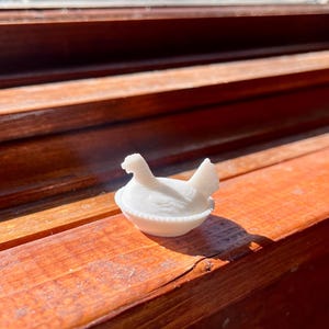 May include: A small, white, opaque glass figurine shaped like a hen sitting on a nest. The base of the nest features a decorative beaded edge, while the hen is depicted with detailed wing feathers and an upright tail, all rendered in solid white glass.