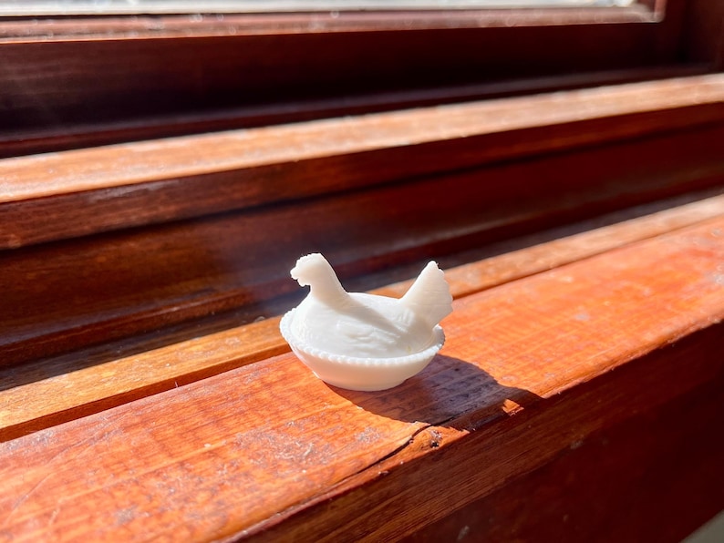 May include: A small, white, opaque glass figurine shaped like a hen sitting on a nest. The base of the nest features a decorative beaded edge, while the hen is depicted with detailed wing feathers and an upright tail, all rendered in solid white glass.