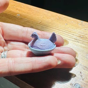 May include: A small, light purple 3D-printed figurine of a chicken sitting inside a bowl. The chicken has a textured surface with visible layer lines from the printing process, a defined head, and a tail. The bowl is a matching light purple color.