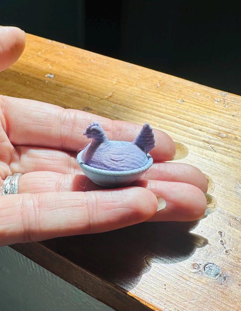 May include: A small, light purple 3D-printed figurine of a chicken sitting inside a bowl. The chicken has a textured surface with visible layer lines from the printing process, a defined head, and a tail. The bowl is a matching light purple color.