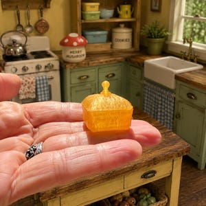 K&ouml;nnte beinhalten: Eine orangefarbene, durchscheinende Miniatur-Butterdose mit quadratischem Boden und passendem Deckel, der oben einen kleinen, spitzen Griff aufweist. Das Objekt besteht aus einem strukturierten, halbtransparenten Material, das das Licht einf&auml;ngt und ihm in einer kleinen K&uuml;chenumgebung ein lebendiges, leuchtendes Aussehen verleiht.