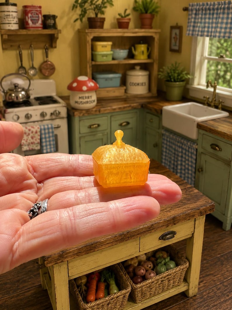 K&ouml;nnte beinhalten: Eine orangefarbene, durchscheinende Miniatur-Butterdose mit quadratischem Boden und passendem Deckel, der oben einen kleinen, spitzen Griff aufweist. Das Objekt besteht aus einem strukturierten, halbtransparenten Material, das das Licht einf&auml;ngt und ihm in einer kleinen K&uuml;chenumgebung ein lebendiges, leuchtendes Aussehen verleiht.