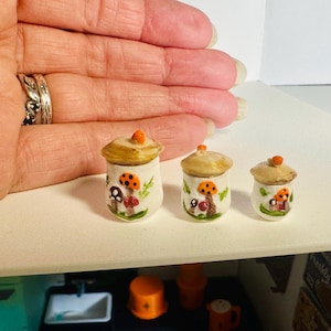 May include: Three miniature white ceramic jars with lids, each featuring hand-painted orange and brown mushroom designs. The jars are arranged in a row from largest to smallest, showcasing a cylindrical shape with a rounded top and a small orange knob.