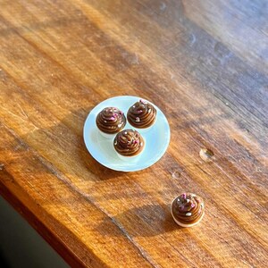 May include: A small white plate holds three miniature chocolate cupcakes with brown frosting and pink sprinkles. One additional cupcake sits separately. The cupcakes are on a wooden surface.