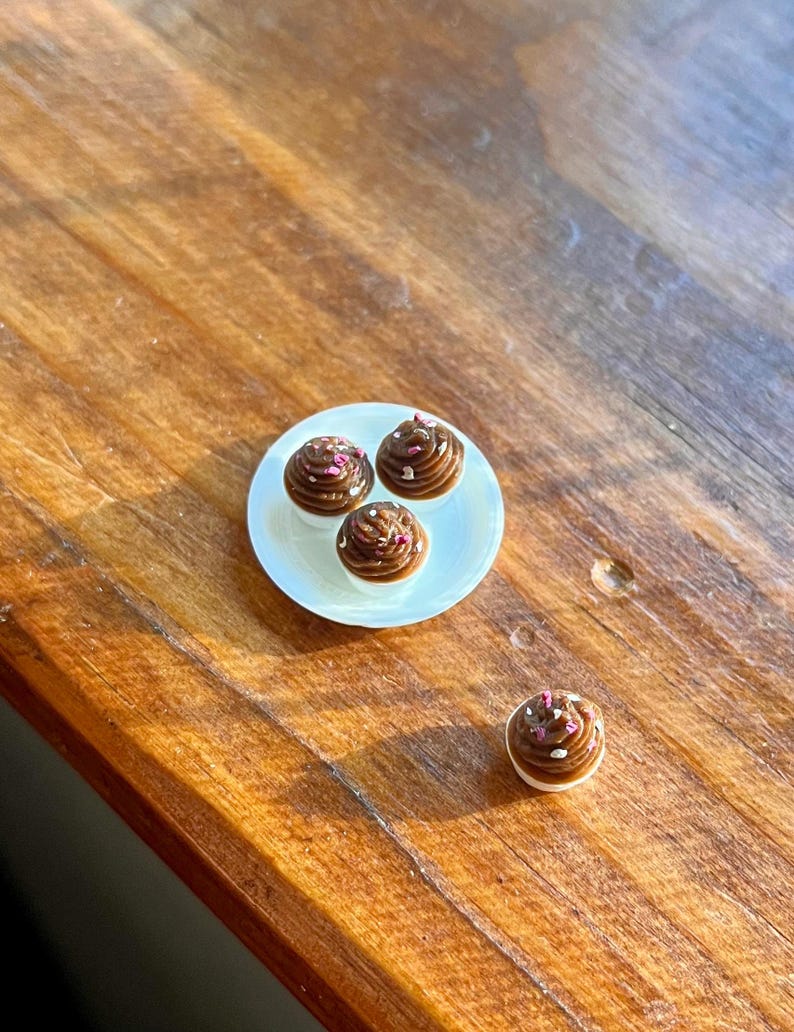 May include: A small white plate holds three miniature chocolate cupcakes with brown frosting and pink sprinkles. One additional cupcake sits separately. The cupcakes are on a wooden surface.